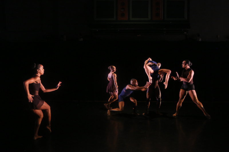 Dancers in blue and rust colored short unitards. A group of dancers make a tableau with the central figure lifted into the air. A single figure stands in the foreground looking towards the group.